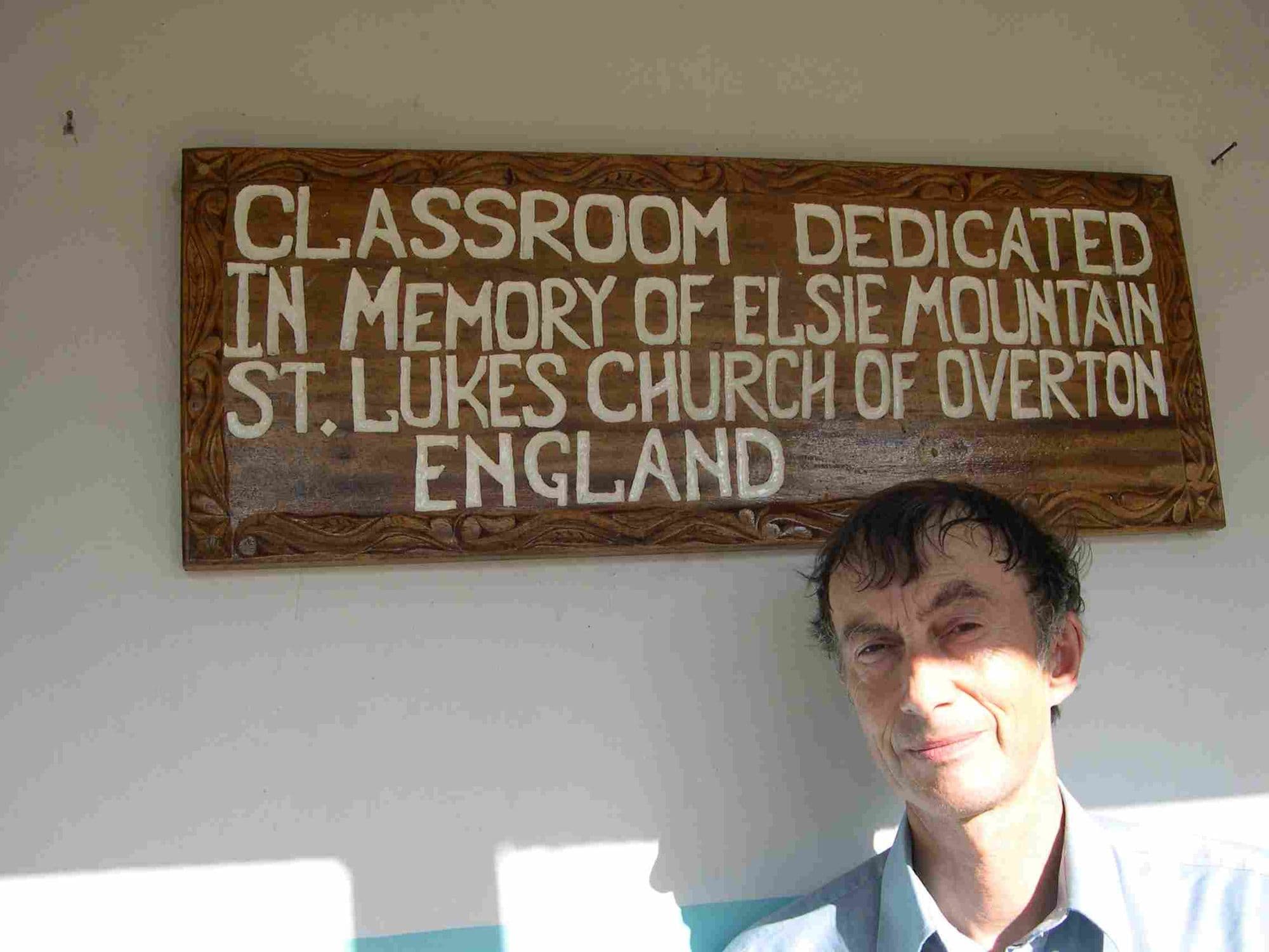 A man beside a carved sign dedicating a classroom to the memory of Elsie Mountain of St Luke's Church, Overton
