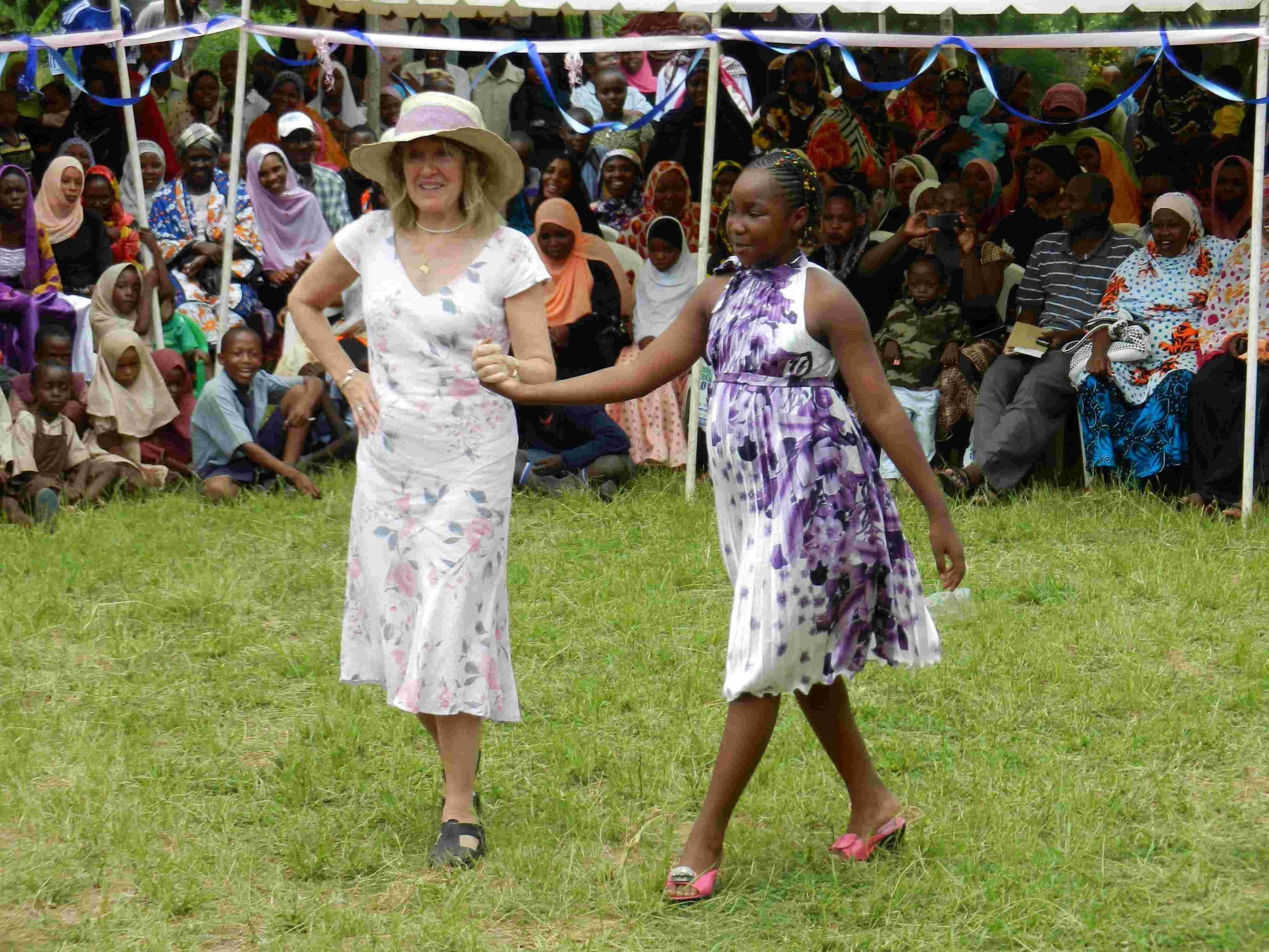 A visitor in a sun hat and a girl in a party dress walking in a fashion show before a crowd
