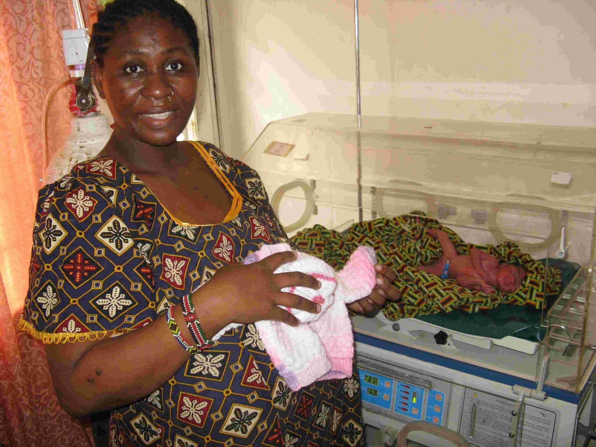 A nurse holding a knitted baby cardigan beside an incubator with a premature baby