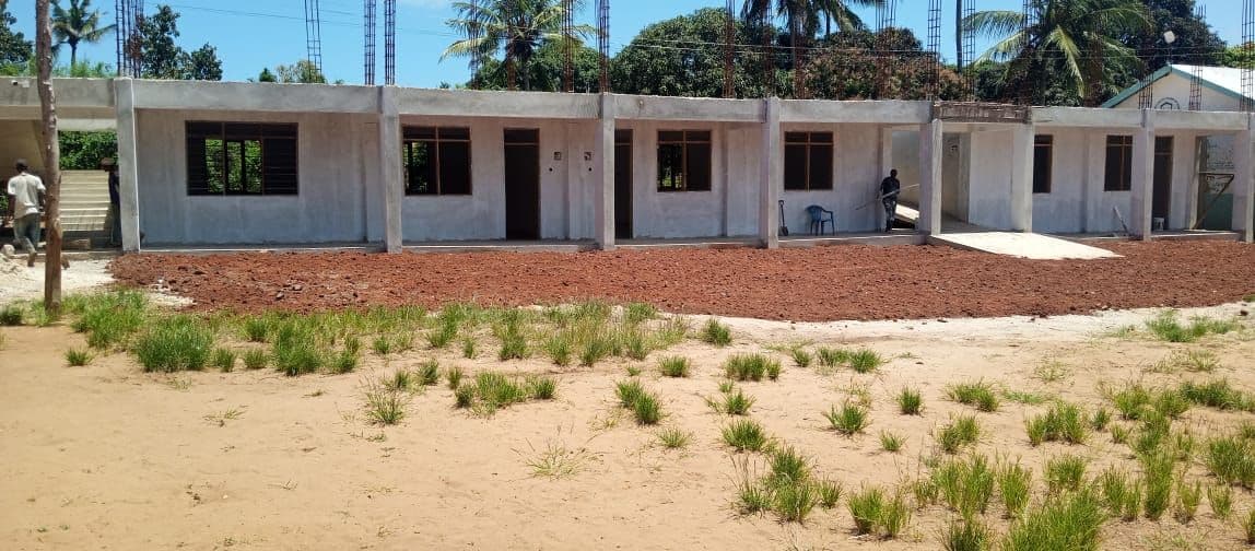 A long row of white single-storey classrooms with a red earth yard and grass in front