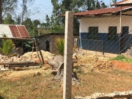 Building with blue-painted lower walls and a rusty iron roof behind a wire fence, with sand and rubble piles