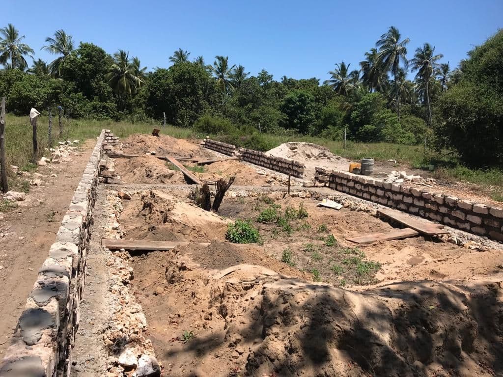 Coral-block foundation walls laid out in sandy ground, with palm trees behind