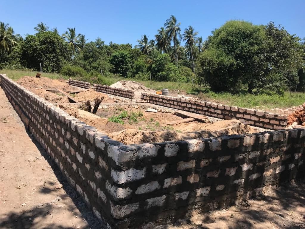 Long coral-block foundation wall of a new building, with sand heaps and palm trees under a blue sky