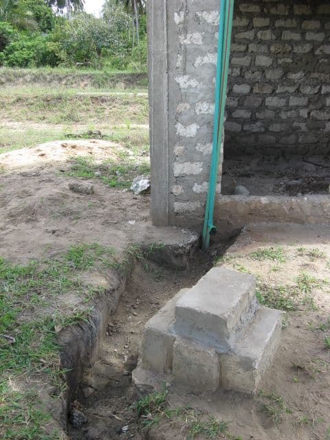 Green water pipe running down a stone building's corner into a trench beside a concrete block