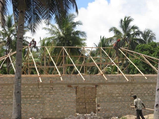 Workers on top of a coral-block wall fixing timber roof rafters among palm trees