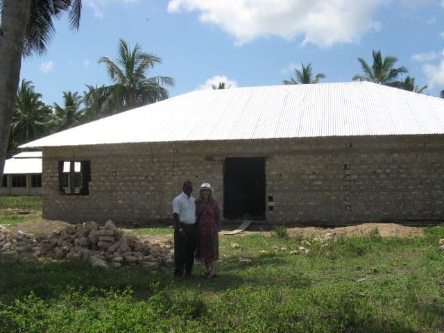Two people standing on grass in front of a coral-block building with a new corrugated iron roof