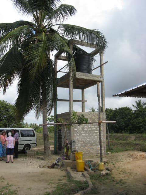 Black water tank on a tall concrete tower beside a palm tree, with yellow jerrycans at a tap below