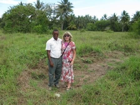 Two people standing on the overgrown, empty plot of land bought for the orphanage, with palm trees behind