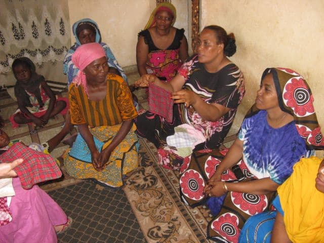 Women seated on a patterned floor mat holding up checked fabric pads during a group session