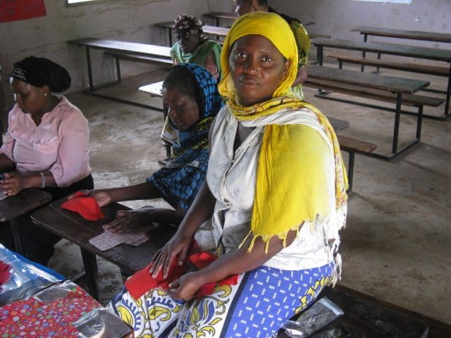 A woman in a yellow headscarf sits at a school desk with red fabric pieces while others sew