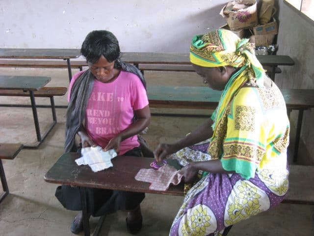Two women at a wooden school desk sewing fabric sanitary pads by hand