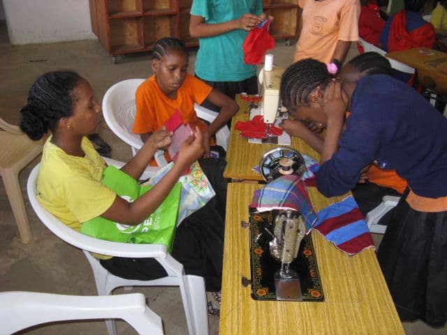 Girls in a classroom sewing fabric pieces on treadle sewing machines