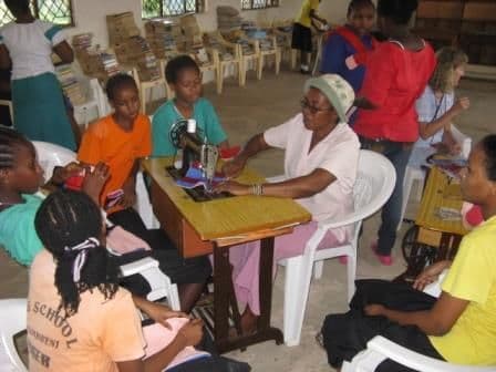 An older woman at a sewing machine helps a group of girls sew in a library