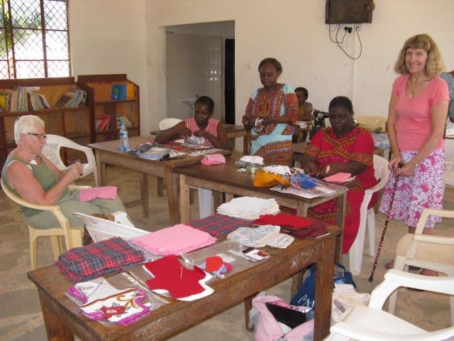 Women at classroom tables sorting fabric pieces and sanitary pad parts with two volunteers